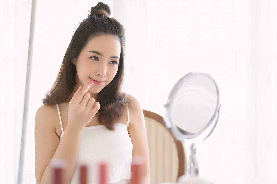 Beautiful Korean girl sitting in front of mirror and applying lipstick on lip in bedroom.
