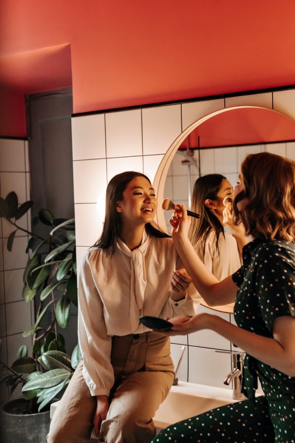 Laughing girls doing makeup together. Curly young woman using cosmetic brush for friend.